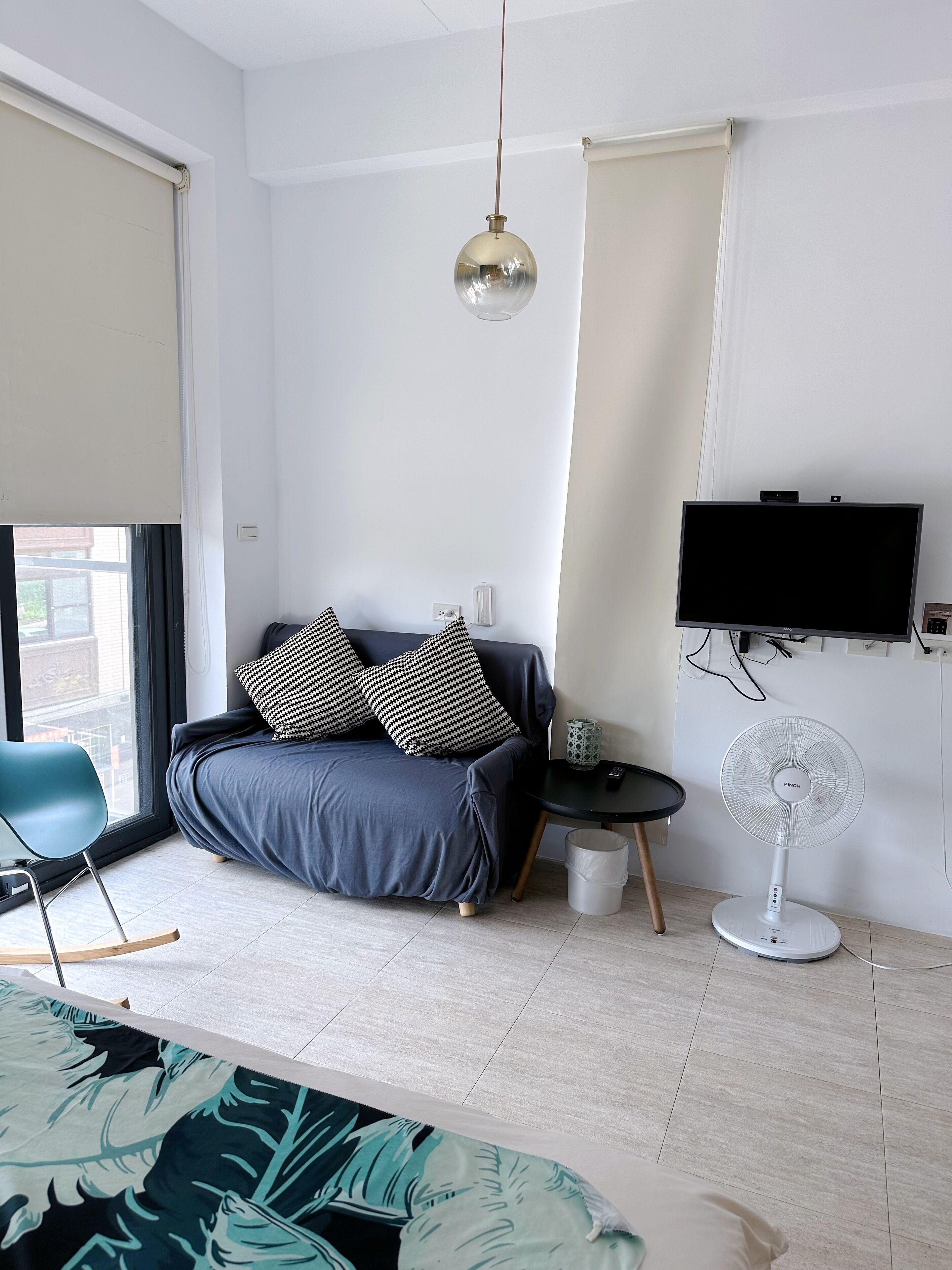 Ein modernes Schlafzimmer mit grauem Sofa, Kissen in Schwarz-Weiß-Muster, einem runden Tisch mit Ventilator und einem an der Wand montierten Fernseher.
