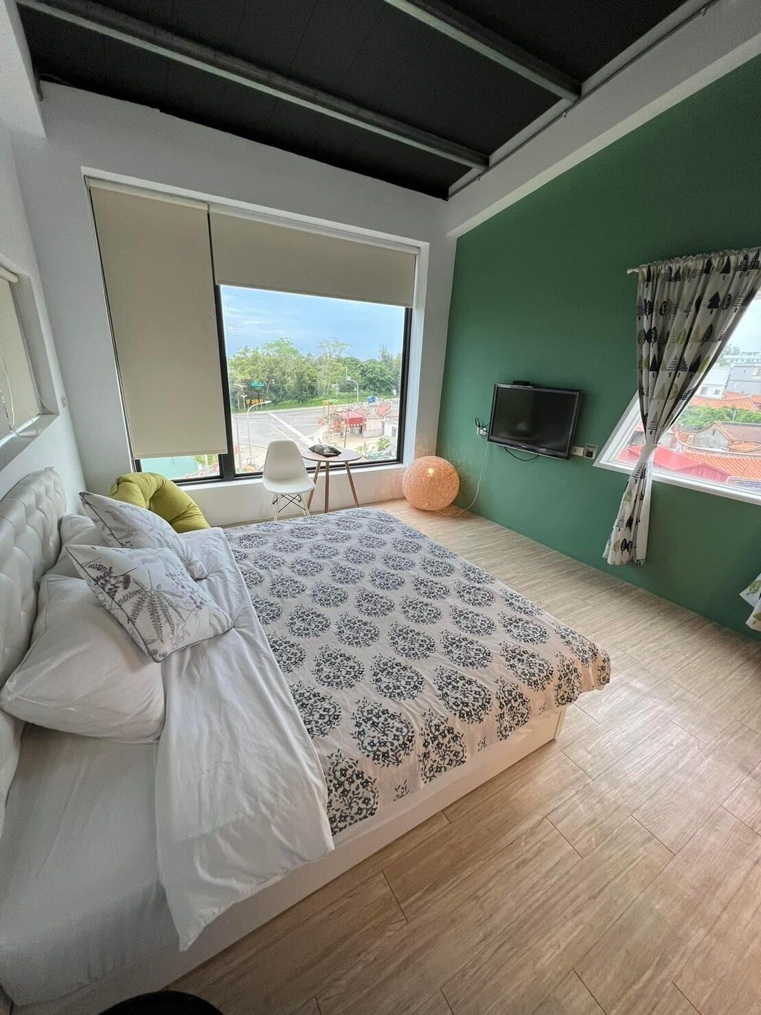 A bedroom with a large bed, a TV mounted on the wall, and a view of a street outside.