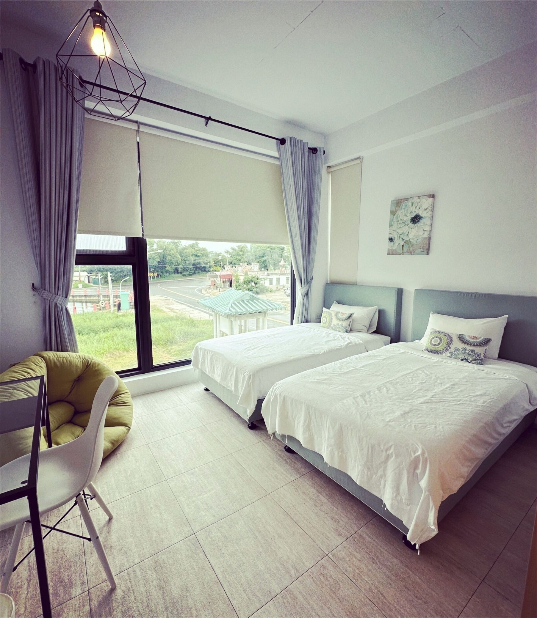 A modern hotel room with two beds, a view of the outside, a dining area with a chair, and a ceiling light fixture.