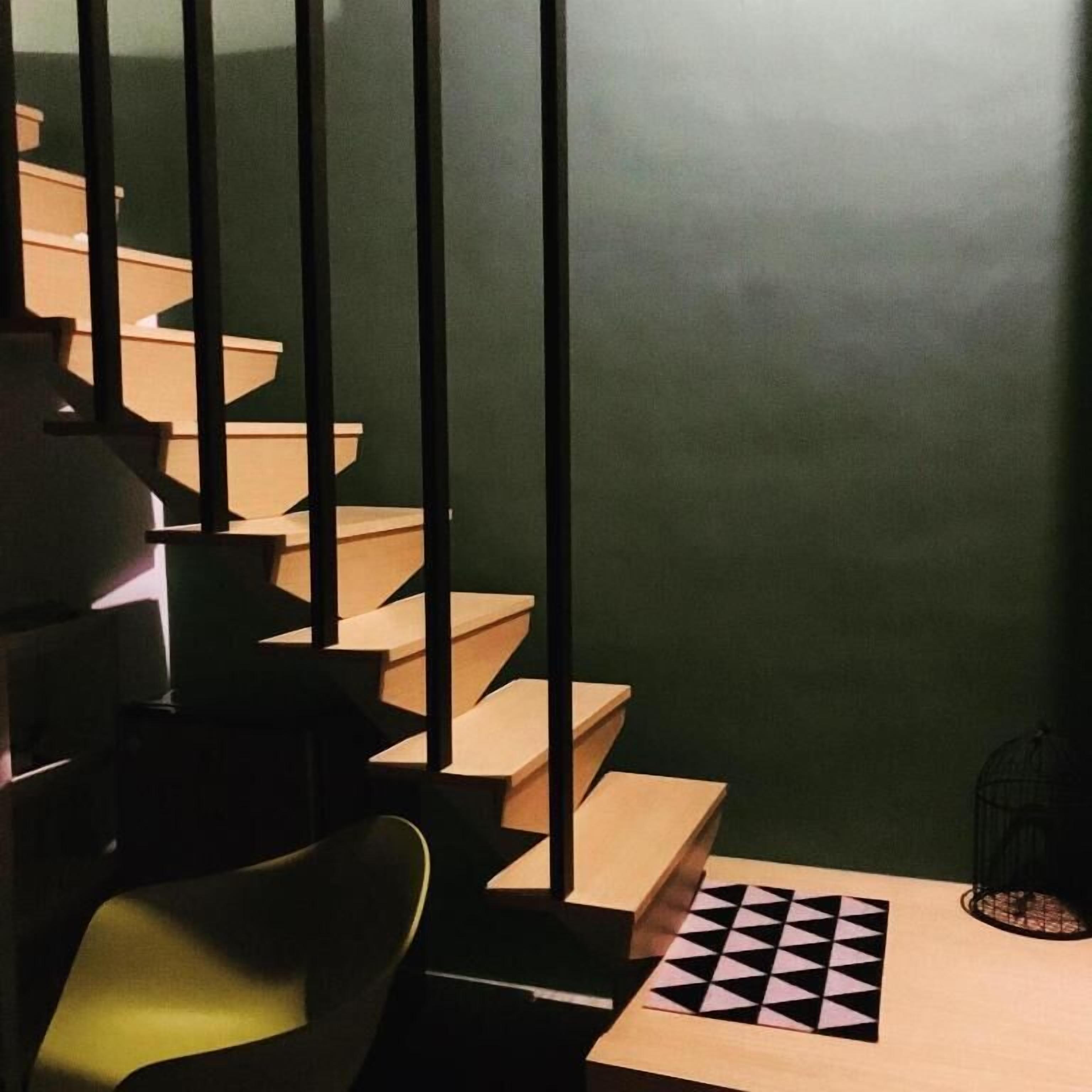 Una escalera con escalones de madera y barandillas negras, que conduce a una habitación con pared verde y un tablero de ajedrez en el suelo.
