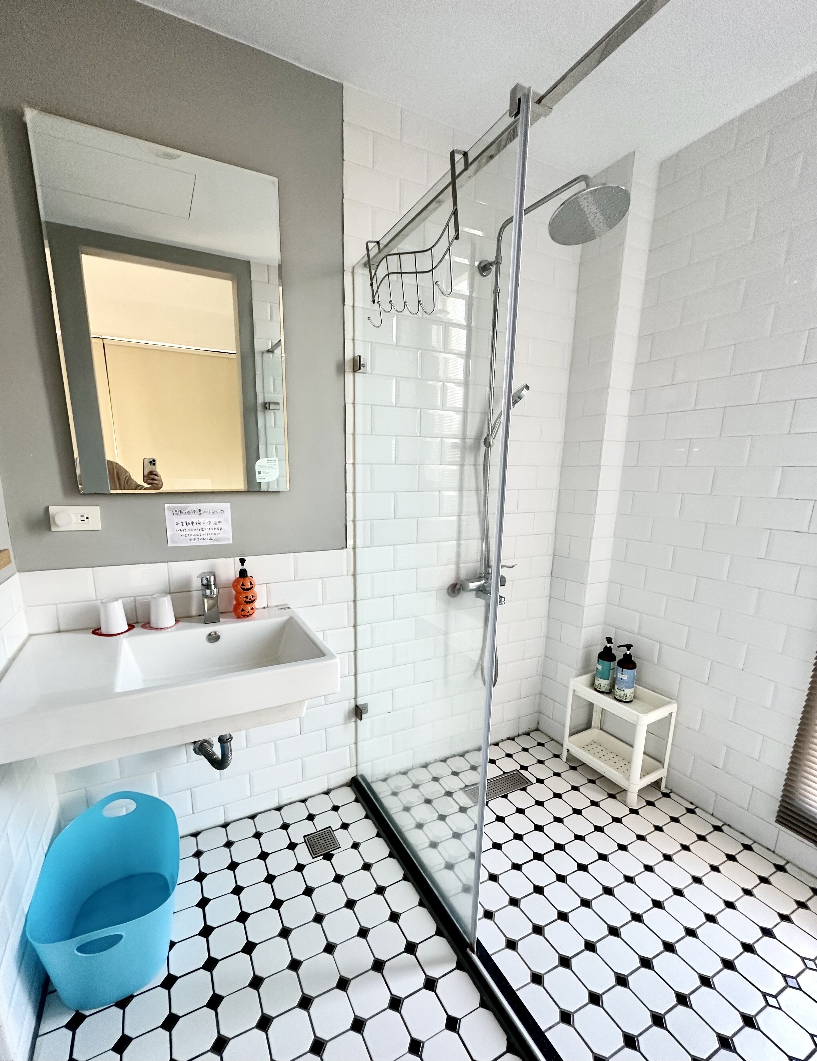 Un baño moderno con ducha de vidrio, un lavamanos blanco con espejo y un estante pequeño con artículos de aseo.
