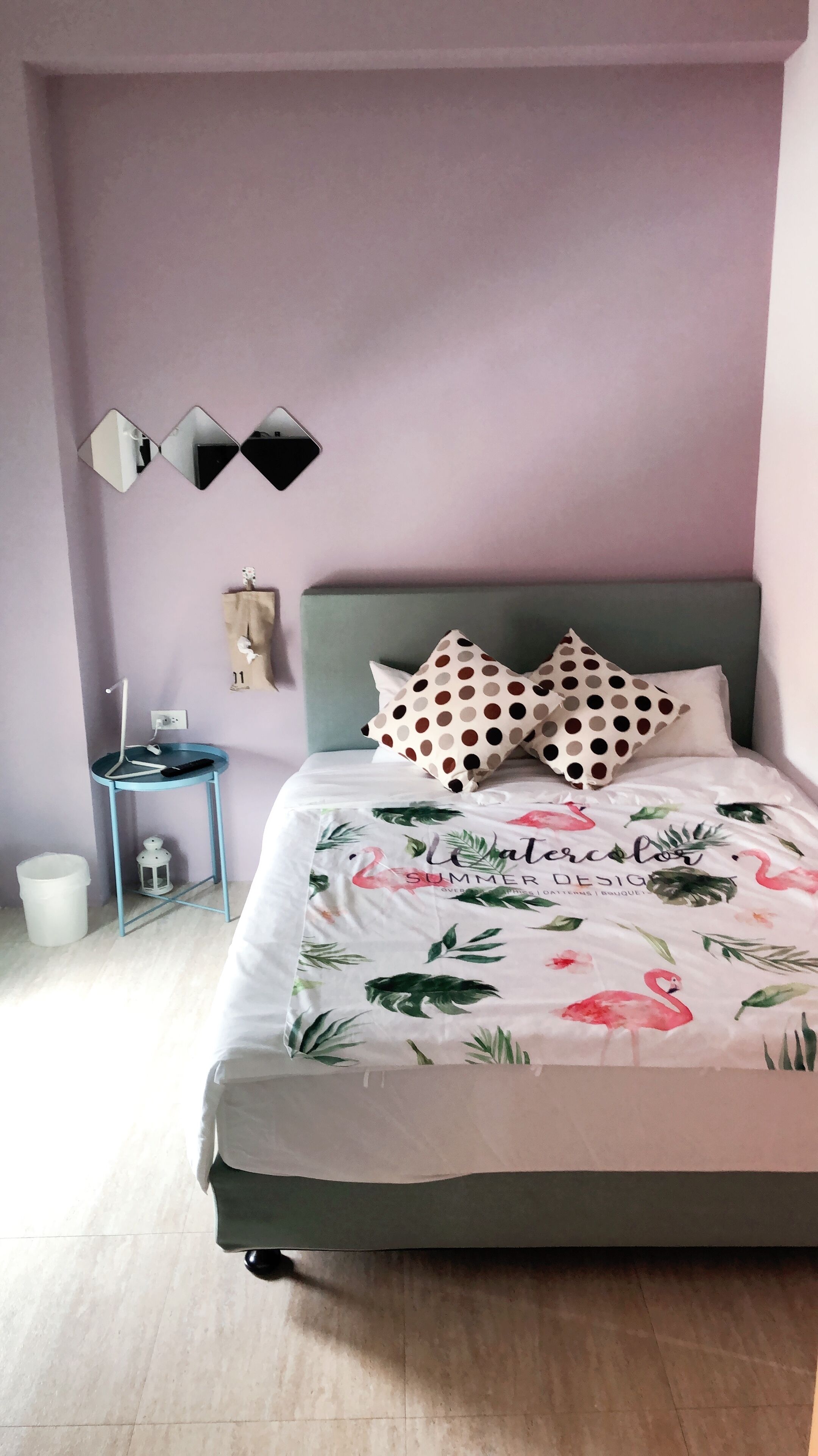 A bedroom with a bed, pillows, a bedside table, a wall-mounted mirror, and a small shelf.