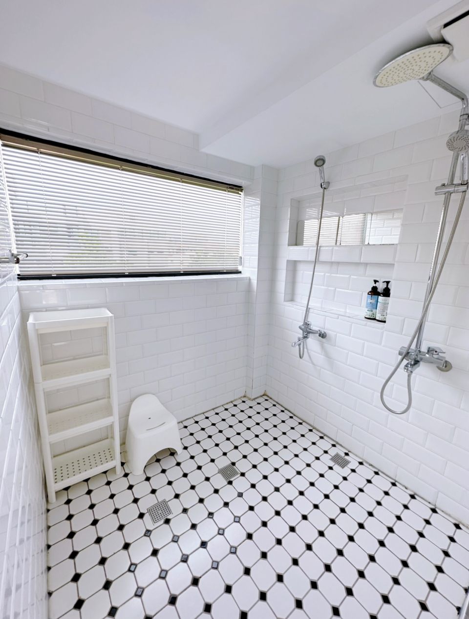 Un baño con paredes y suelo de azulejos blancos, ducha con cabezal de ducha manual y ventana con persianas.