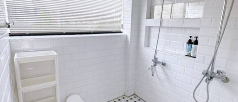 Un baño con paredes y suelo de azulejos blancos, ducha con cabezal de ducha manual y ventana con persianas.