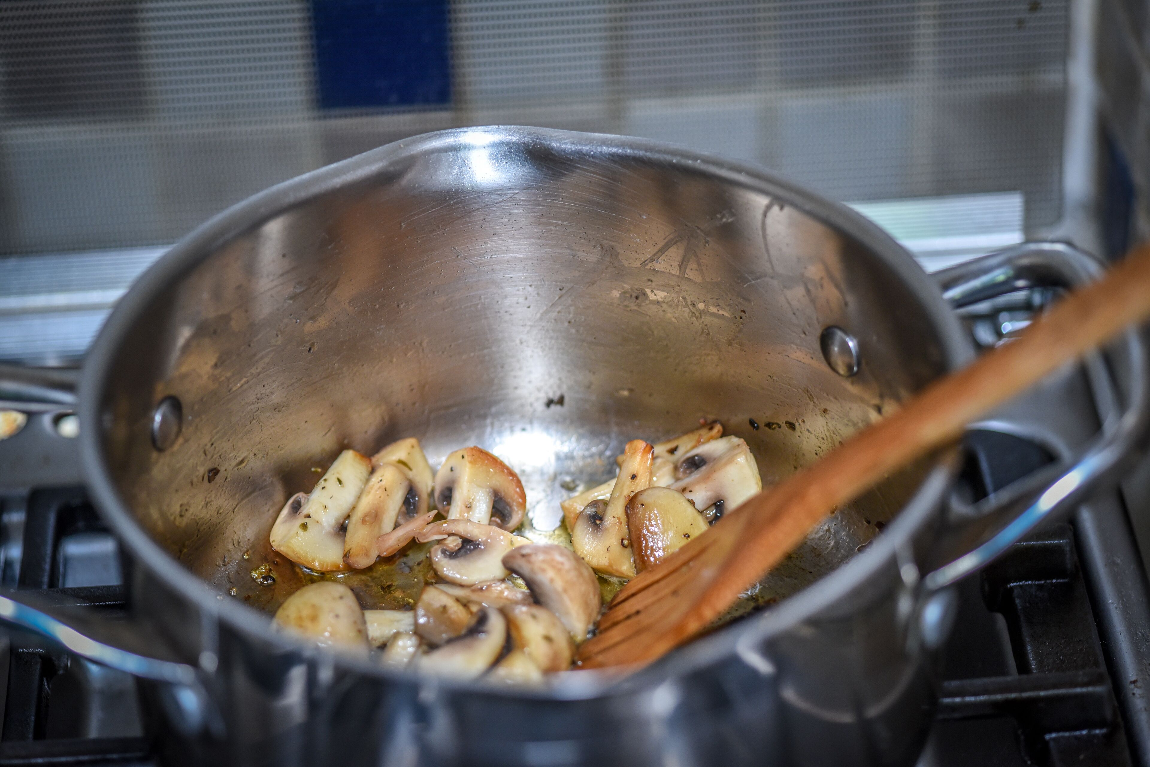 Une poêle en acier inoxydable sur une cuisinière, avec des champignons coupés en tranches et une spatule en bois.