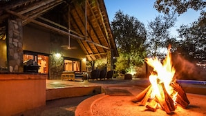 A fire pit with logs, a stone structure, and a building with a sloped roof in the background. - Leopard's Lair Bush Lodge (Hoedspruit)