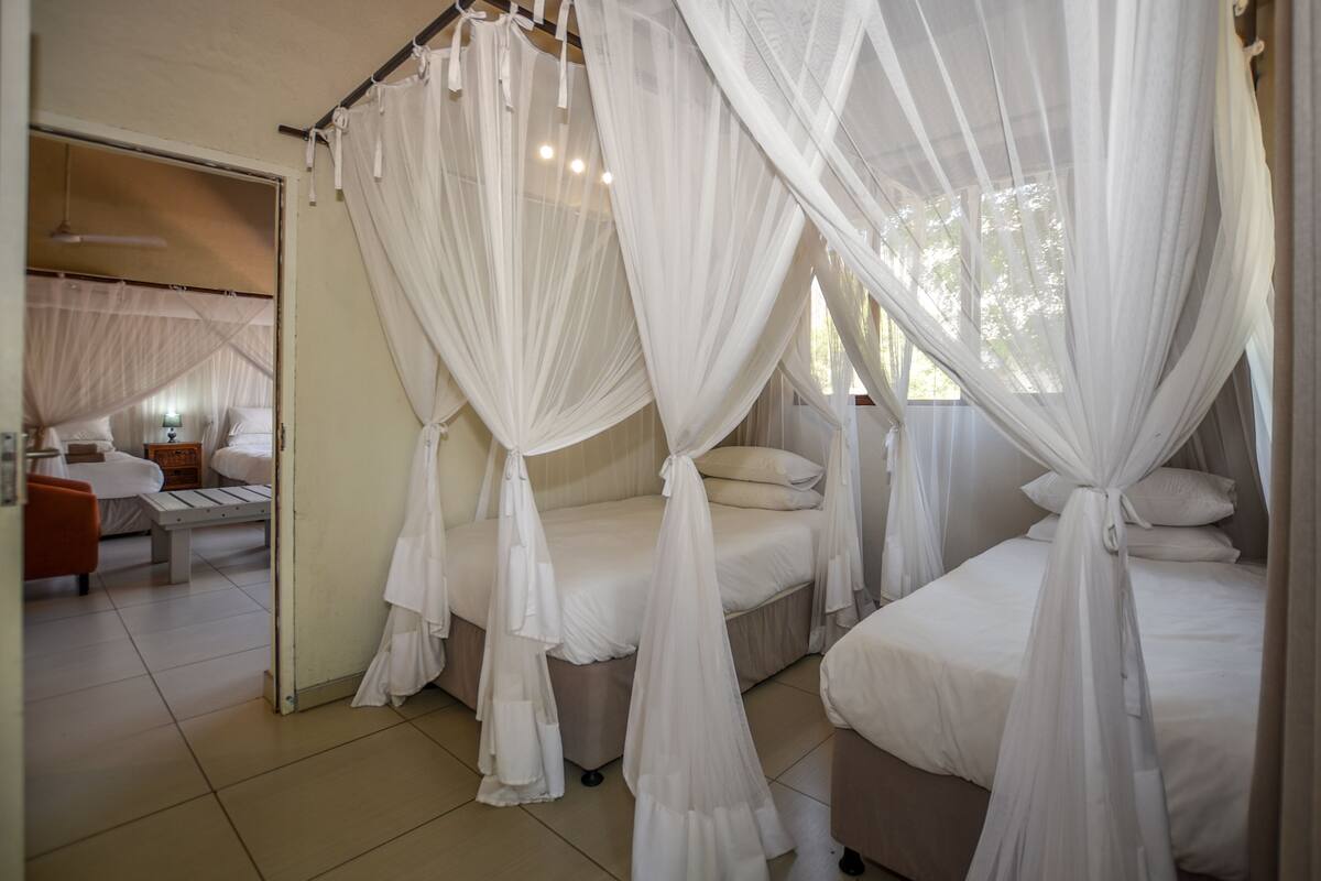 A room with three beds, each covered with white canopy beds. There is a bench and a table with chairs in the corner.