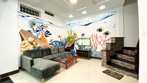 Una sala de estar con un sofá seccional gris, una mesa de centro de vidrio, un televisor de pantalla plana y un mural en la pared que muestra una escena submarina.