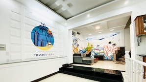 Vestíbulo de hotel con diseño moderno, que presenta un gran mural en la pared, un área de descanso con sofá y televisor, y una escalera con barandilla blanca.