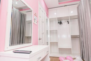 Une salle de bain rose et blanche avec un meuble-lavabo, un miroir, un rideau de douche et une armoire blanche où sont suspendus des vêtements.