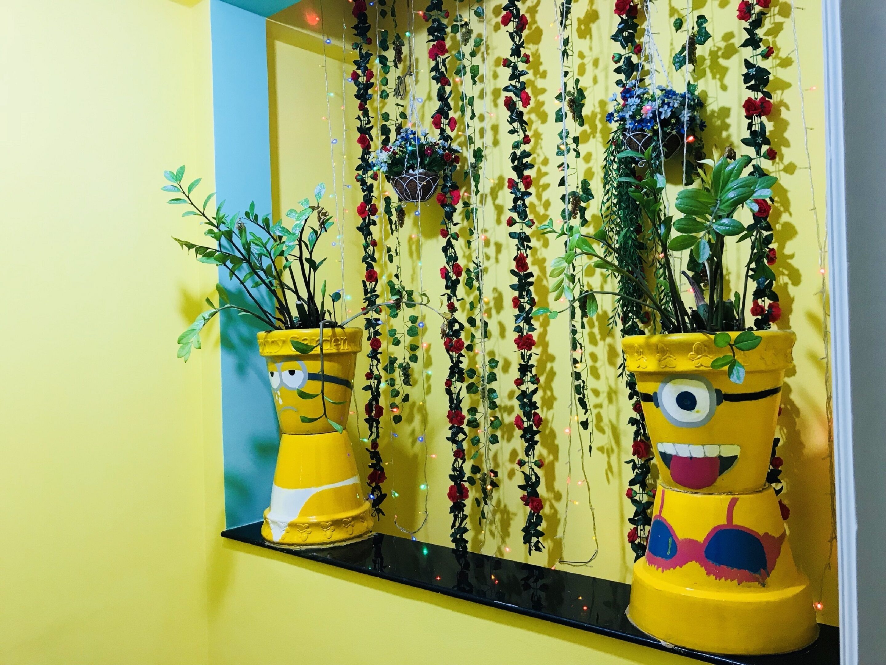 A yellow wall with a mirror reflecting a window sill decorated with potted plants and string lights.