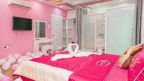 Een hotelkamer in roze tinten met een bed, een televisie, een douche en een kaptafel.