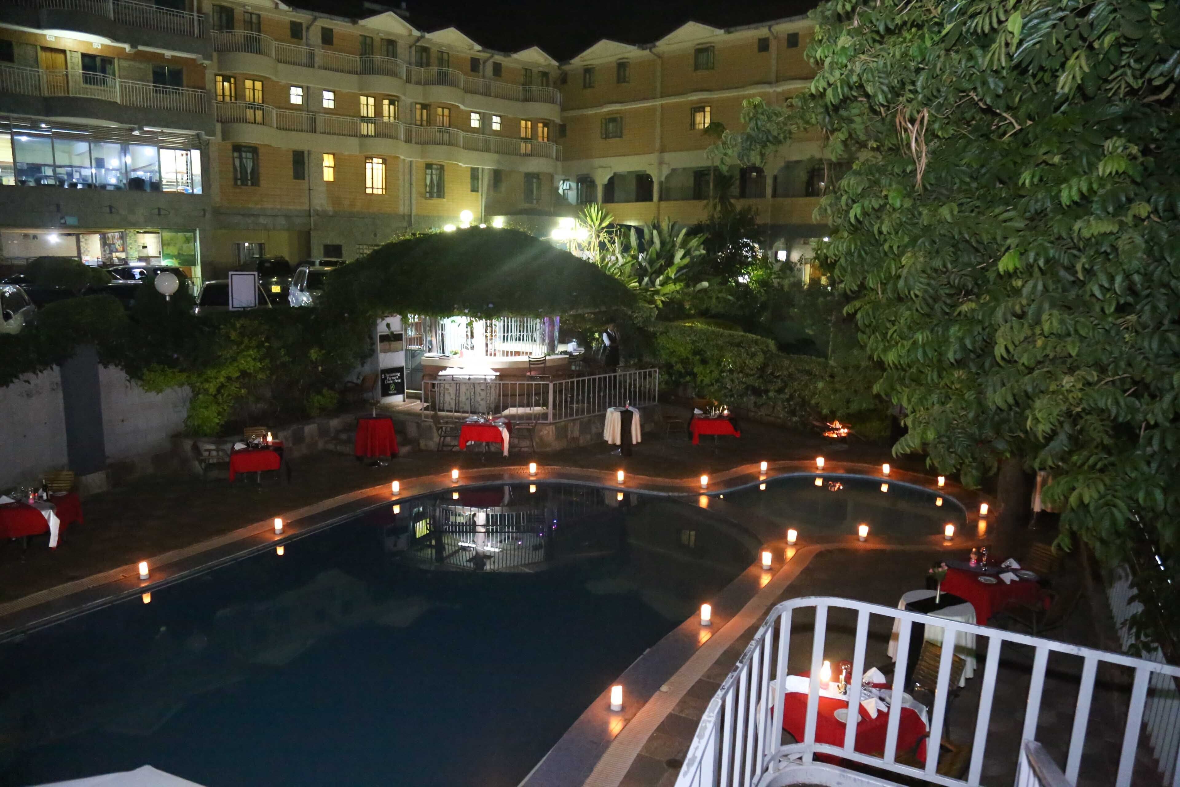 An outdoor swimming pool with a surrounding deck, red tables, and a bar area with a thatched roof.