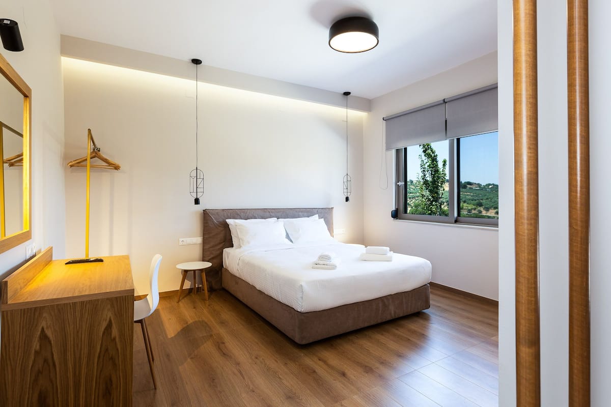 A modern bedroom with a wooden desk, a hanging light fixture, a bedside table, and a large window with a view of greenery.
