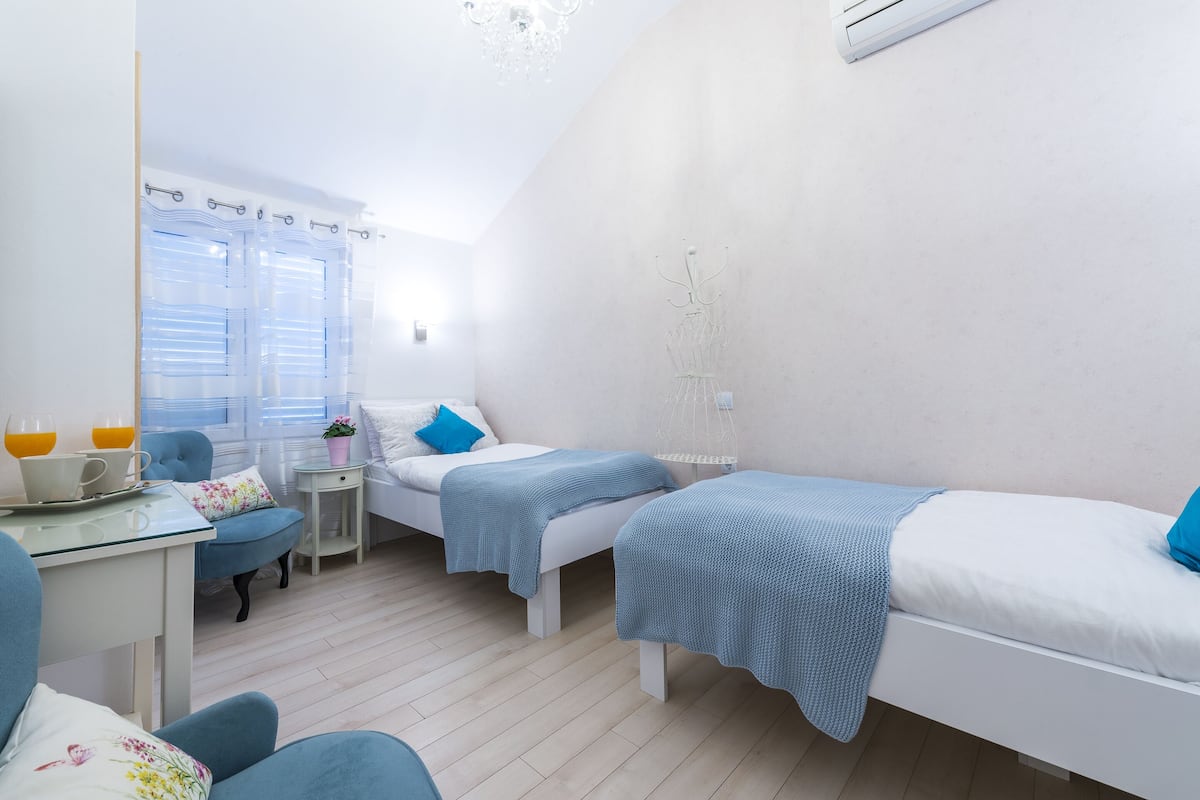 A small, well-lit room with two beds, a desk with a chair, and a window with curtains.