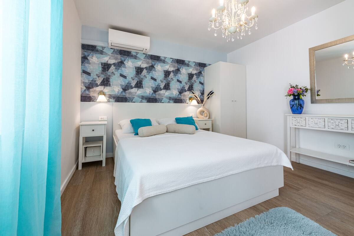 A modern bedroom with a large bed, white walls, a geometric-patterned accent wall, a chandelier, a bedside table with a lamp, and a blue vase with flowers.