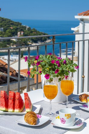 A table with watermelon slices, two glasses of orange juice, a croissant, and a cup on a balcony overlooking a coastal view. - Villa Boro 4 (Dubrovnik)