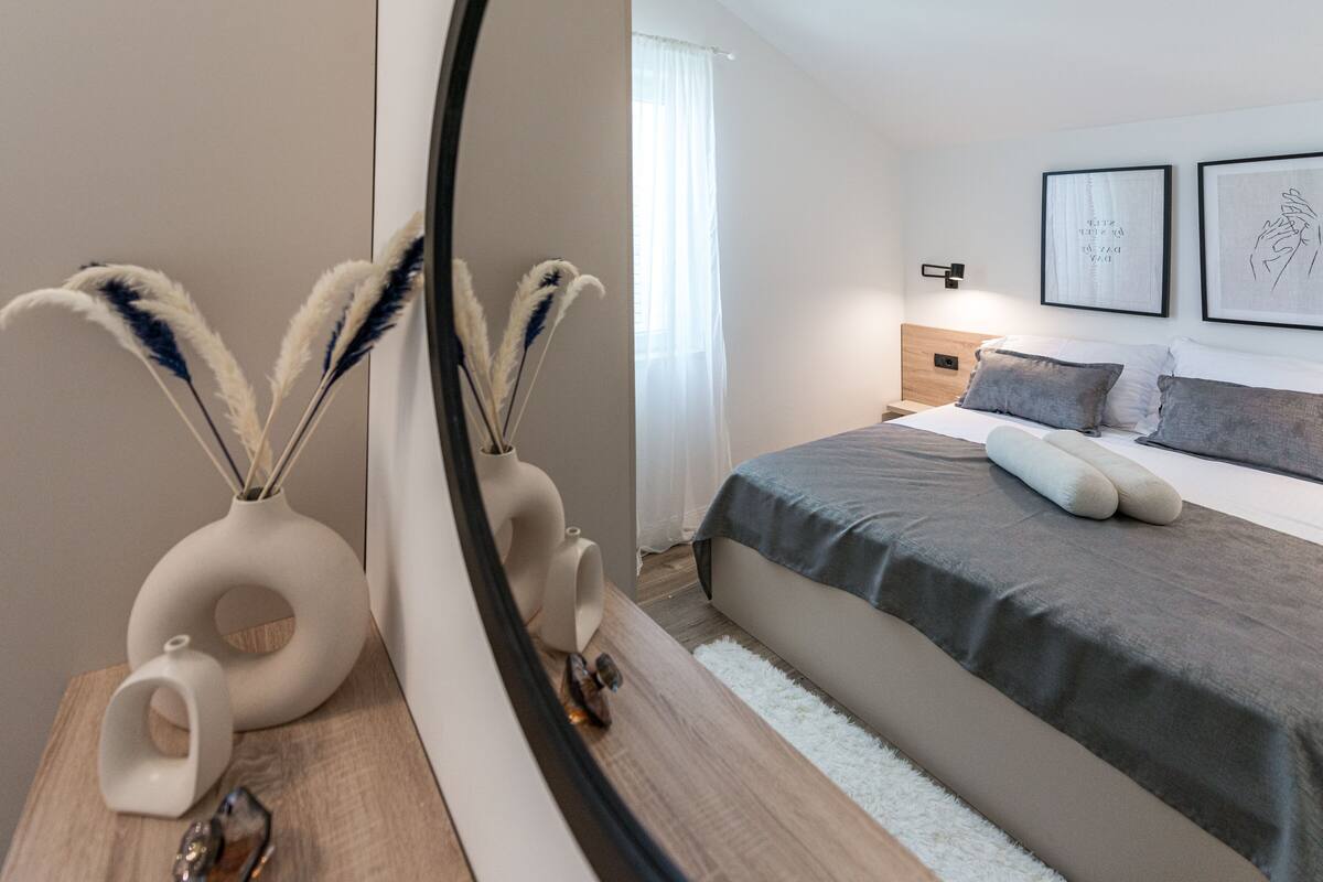 Une chambre à coucher moderne avec un grand miroir, des vases décoratifs, une table de chevet en bois, un lit avec une literie grise et deux œuvres d’art encadrées accrochées au mur.