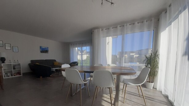 A spacious living area with a dining table, chairs, a sofa, and a large window offering a view of the outdoors. - Beautiful new contemporary house 2 min walk from the beaches of Cabellou (Concarneau)