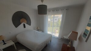A bedroom with a bed, a nightstand, a desk, a chair, a lamp, a hanging fan, a window with curtains, and a view of the outside. - Beautiful new contemporary house 2 min walk from the beaches of Cabellou (Concarneau)