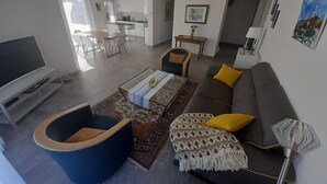 A modern living room with a sofa, armchair, and coffee table. There is a TV, a dining area, and a kitchen in the background. - Beautiful new contemporary house 2 min walk from the beaches of Cabellou (Concarneau)