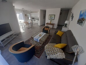A modern living room with a sofa, armchair, and coffee table. There is a TV, a dining area, and a kitchen in the background.