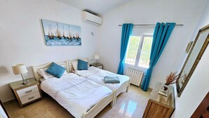 A bedroom with two beds, a painting of sailboats, an air conditioner, a window with curtains, and a mirror. - Villa Bienestar Jávea with pool (Xàbia)