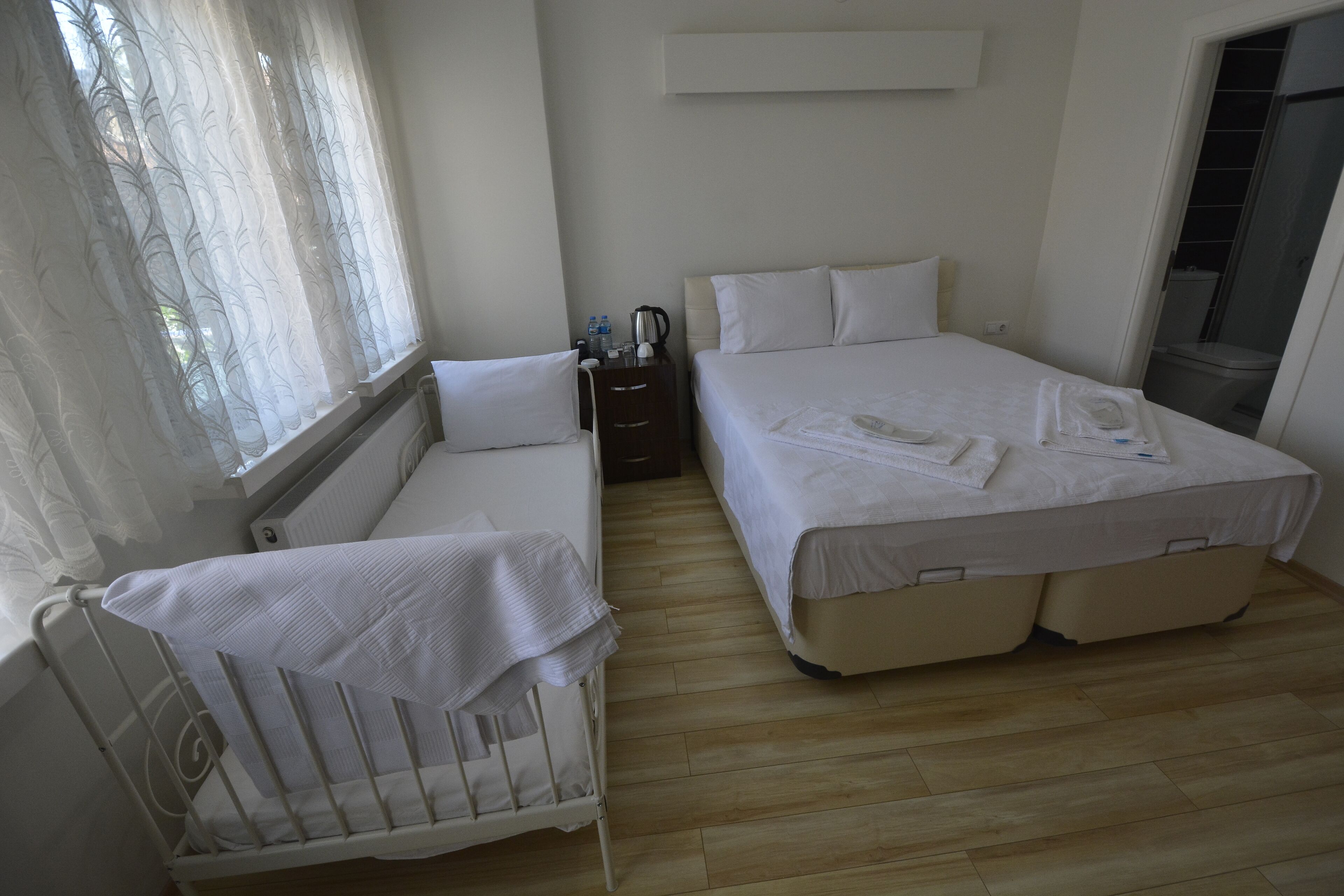 Hotellihuone, jossa on vauvansänky, parivuode, yöpöytä lampulla ja ikkuna, jossa on läpikuultavat verhot.