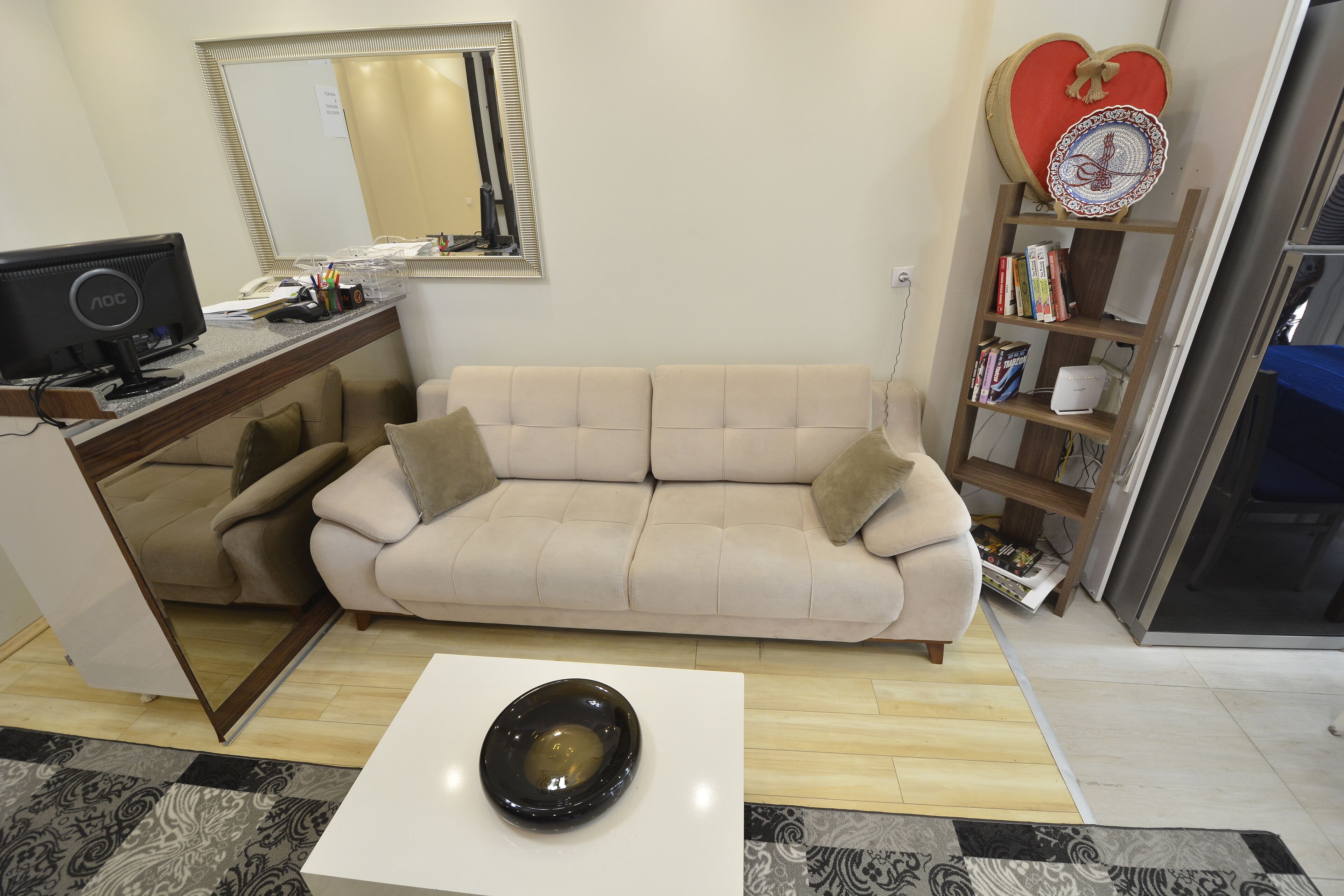 A living room with a beige sofa, a white coffee table with a black bowl, a desk with a computer, a bookshelf, and a heart-shaped decoration on the wall.