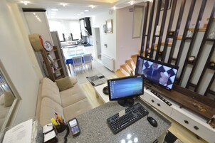 Una sala moderna con un televisor de pantalla plana grande, una computadora y una mesa de centro. Se observa un sofá y un área de comedor al fondo.