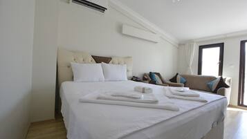 A neatly made bed with white linens and towels, a beige sofa with blue and brown cushions, and a window with curtains.