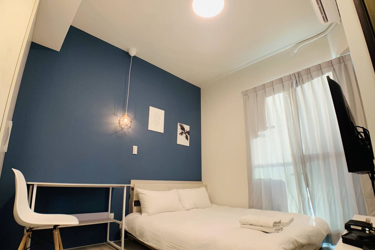 A bedroom with a bed, a desk, a chair, a TV, and a window with curtains.