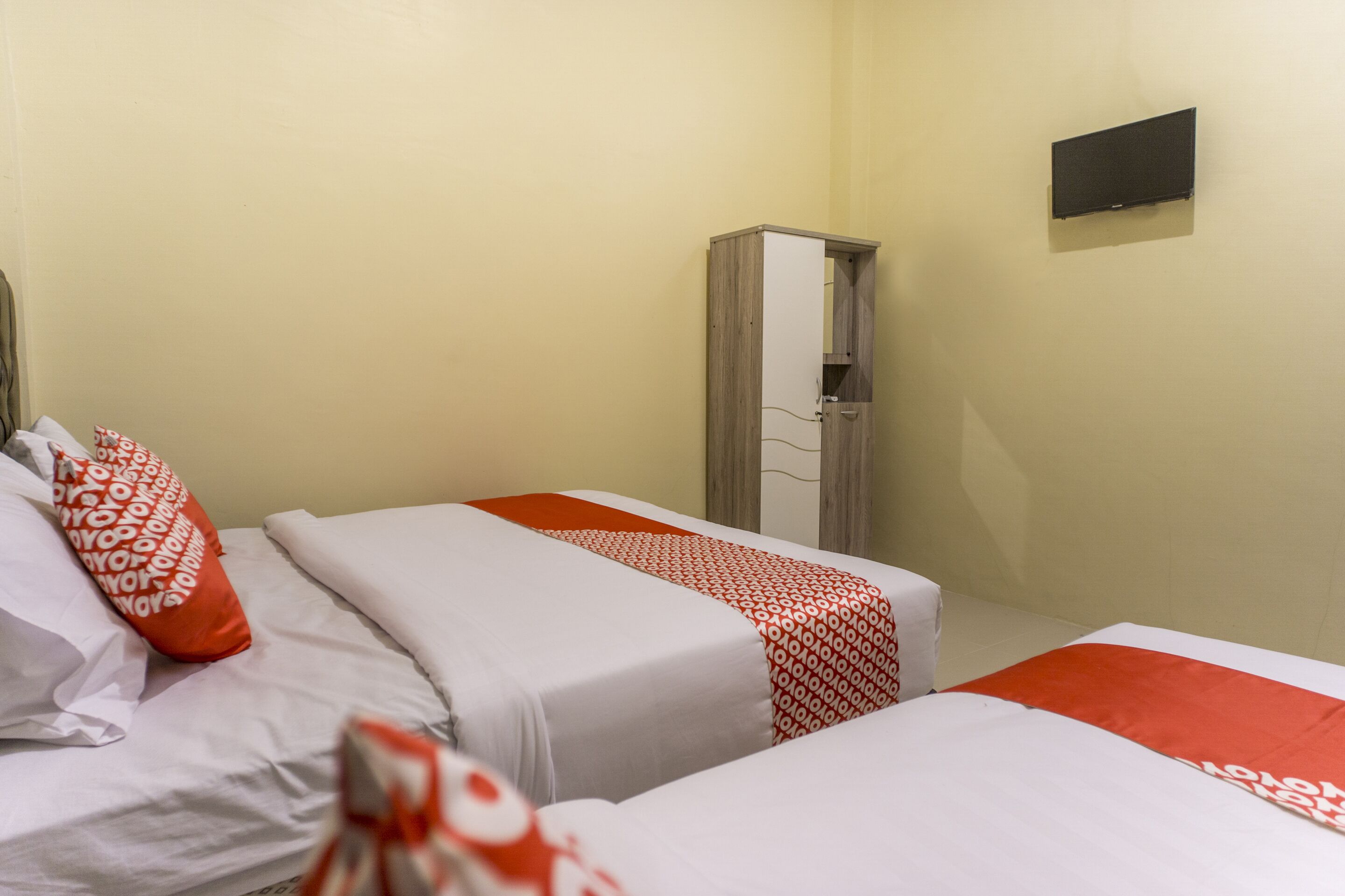 Hotellihuone, jossa on kaksi sänkyä, ja molemmissa on valkoiset ja punaiset vuodevaatteet. Huoneessa on myös vaatekaappi ja seinälle asennettu taulutelevisio.