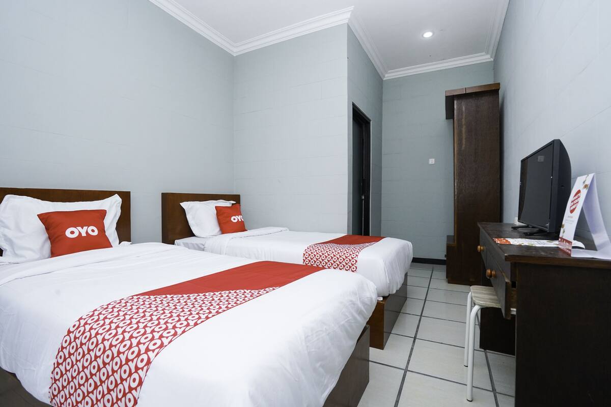 A hotel room with two beds, each with white bedding and red accents, a desk with a computer, and a television.