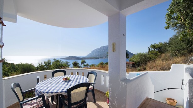 A covered outdoor seating area with a table and chairs, overlooking a scenic view of the sea and a mountain.