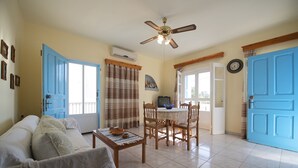 A living room with a sofa, a coffee table, and a dining area with chairs and a table. There is a ceiling fan, a clock on the wall, and a door with a blue frame.