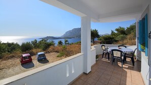 A balcony with a view of the sea, a table with chairs, and two parked cars.