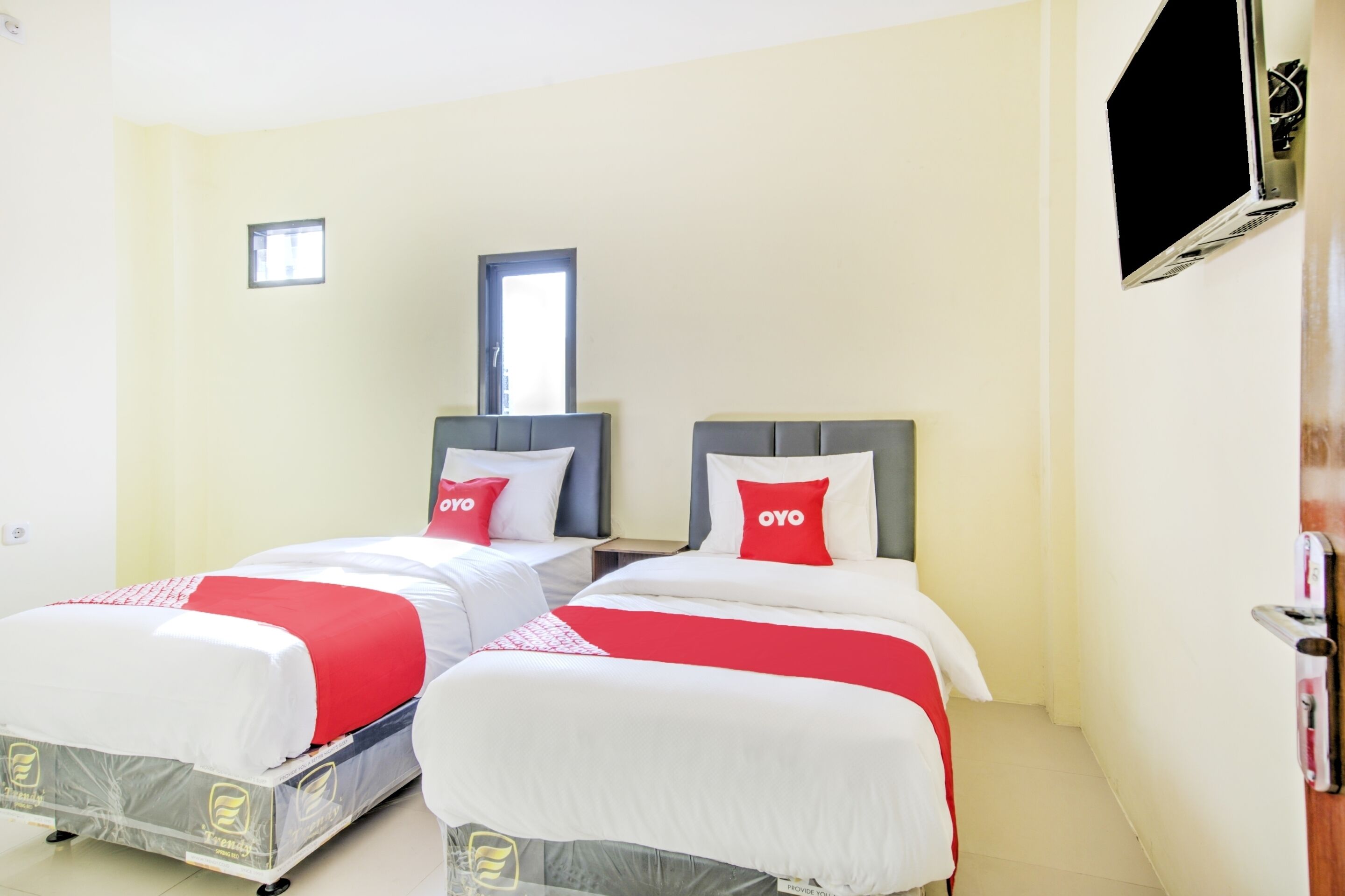 A hotel room with two beds, each with a red and white bedspread and pillows, a small window, and a flat-screen TV mounted on the wall.