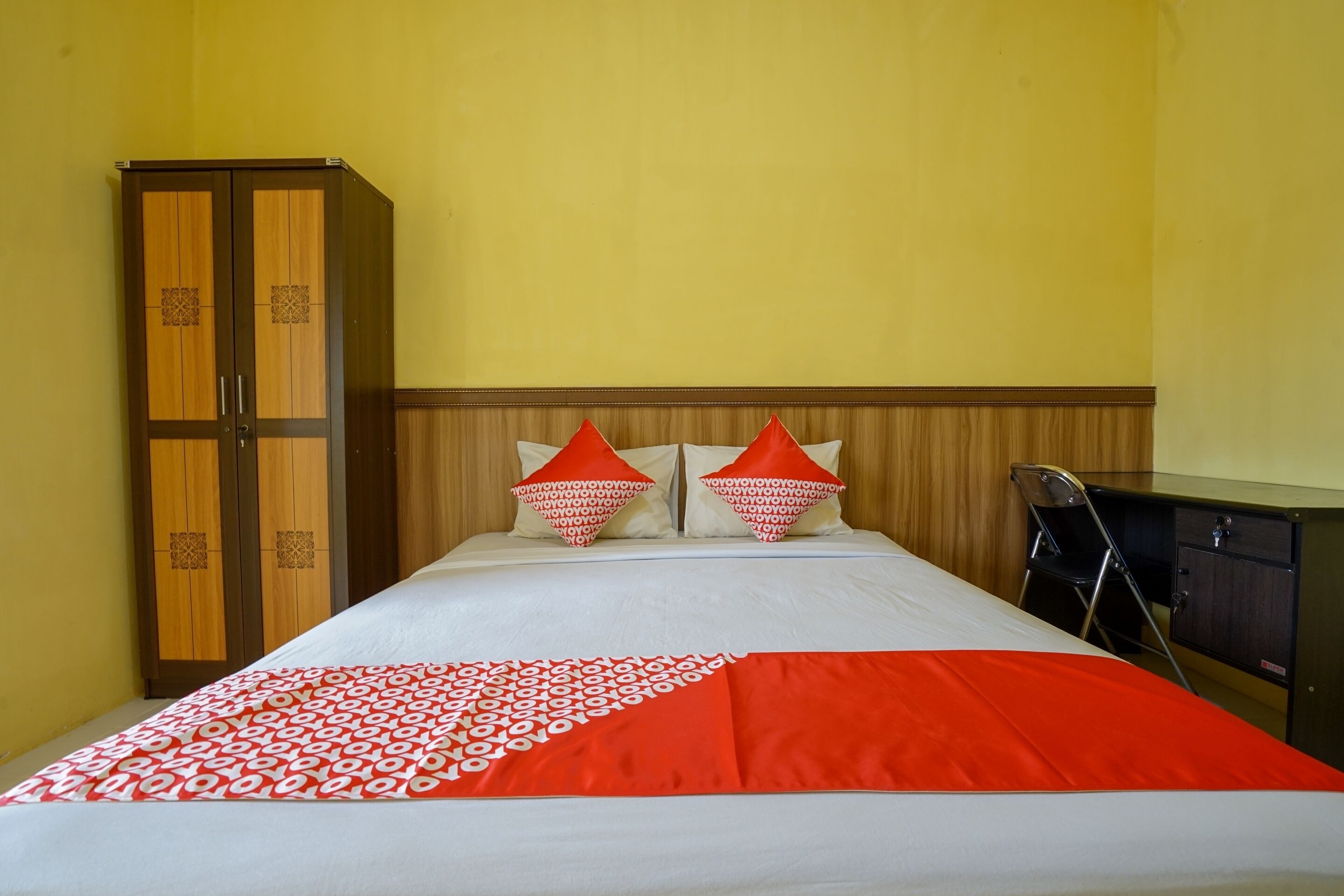 A hotel room with a bed, two pillows, a red and white bedspread, a wooden wardrobe, and a desk with a chair.