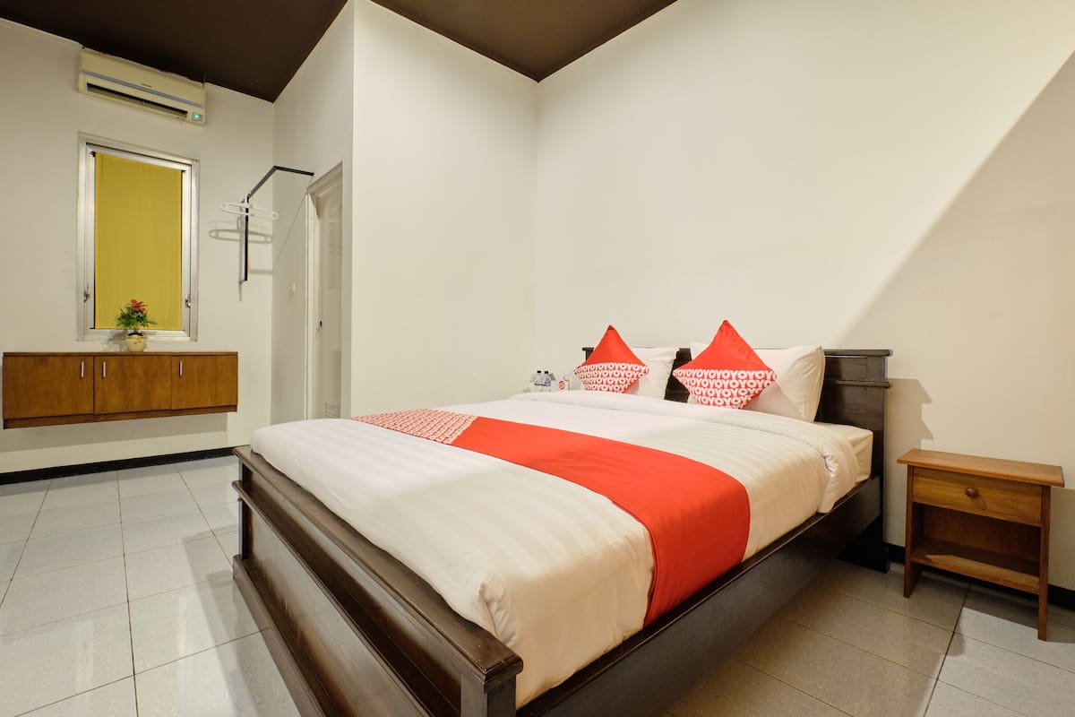 A bedroom with a bed, a wooden nightstand, a small table with a lamp, a window with a curtain, and a wall-mounted air conditioner.