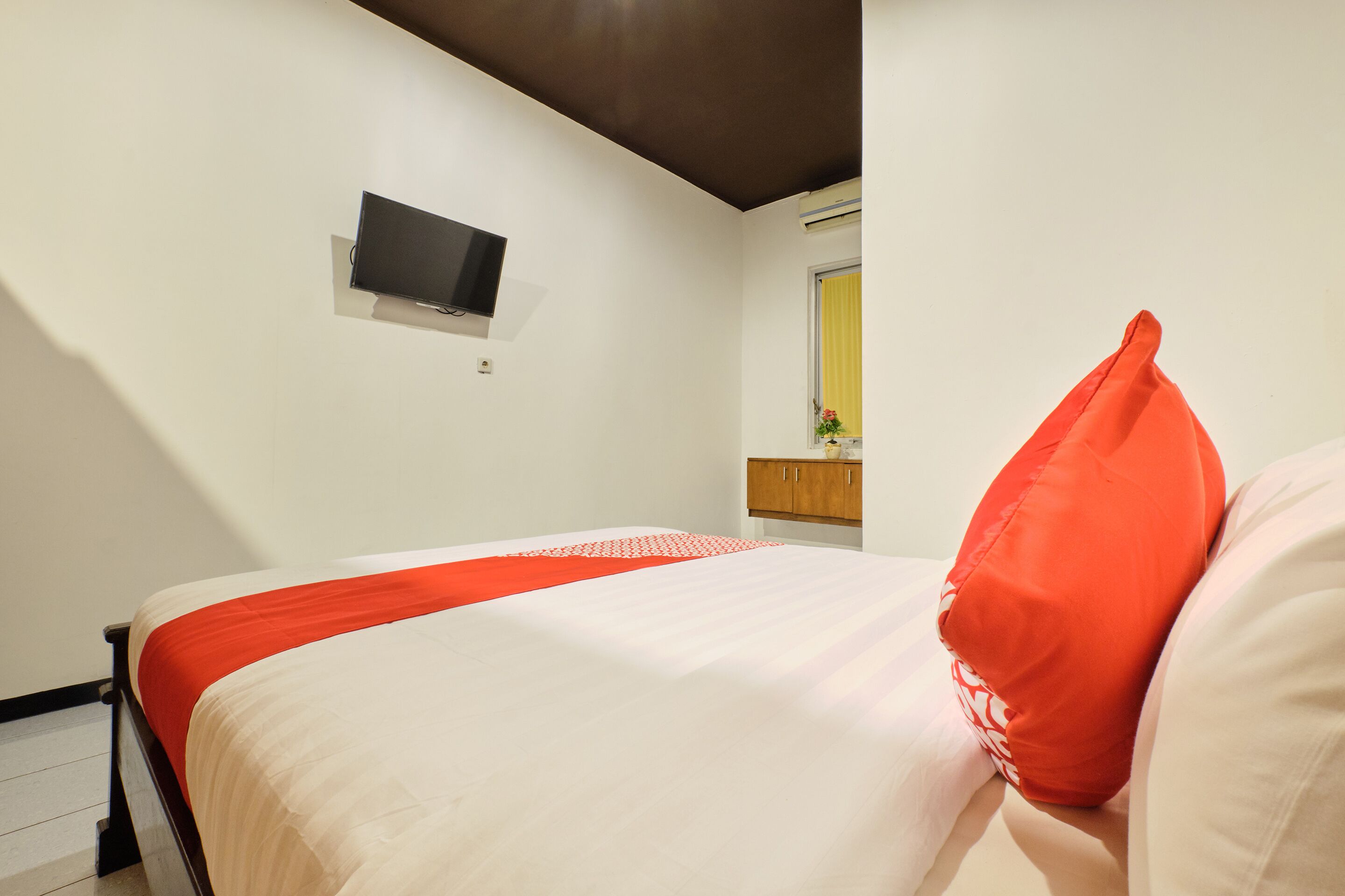 A neatly made bed with a red accent pillow, a mounted television on the wall, and a window with a yellow curtain.