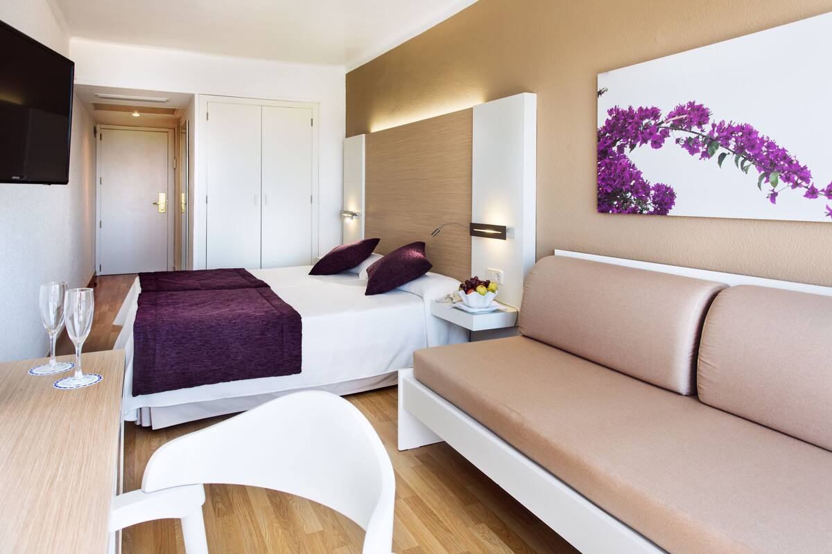 A modern hotel room with a bed, a sofa, a dining table with two glasses, and a wall-mounted TV.