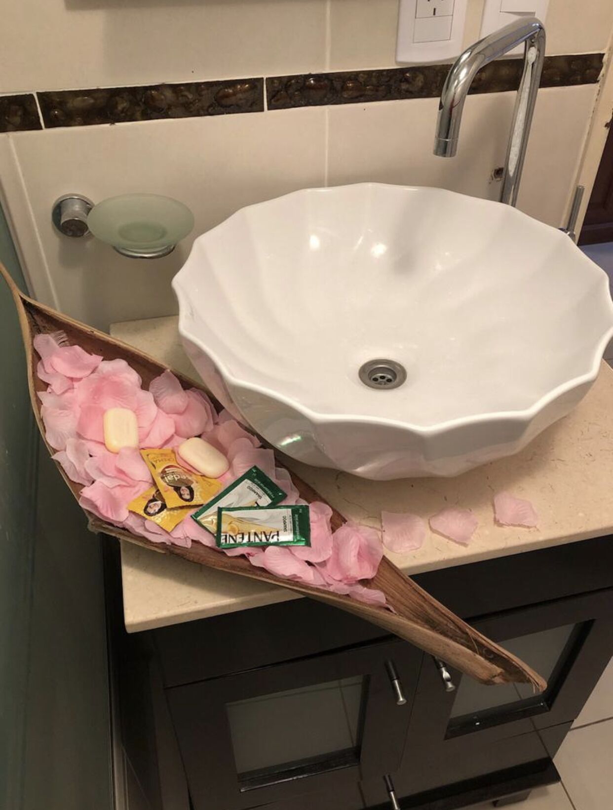 A bathroom with a white sink, a wooden decorative piece with pink petals and toiletries, and a glass soap dish.