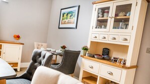 A cozy dining area with a table set for two, a cushioned chair, and a cabinet with glass doors containing kitchenware. - Little Regent Hall (Sheringham)