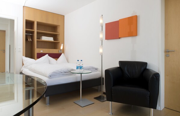 A modern hotel room with a bed, a glass table, a black leather chair, a floor lamp, and a wardrobe with shelves.