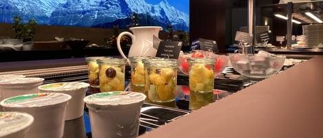 Una zona de bar con una pantalla grande mostrando un paisaje de montaña, varios frascos de ensalada de frutas y una jofaina de agua.