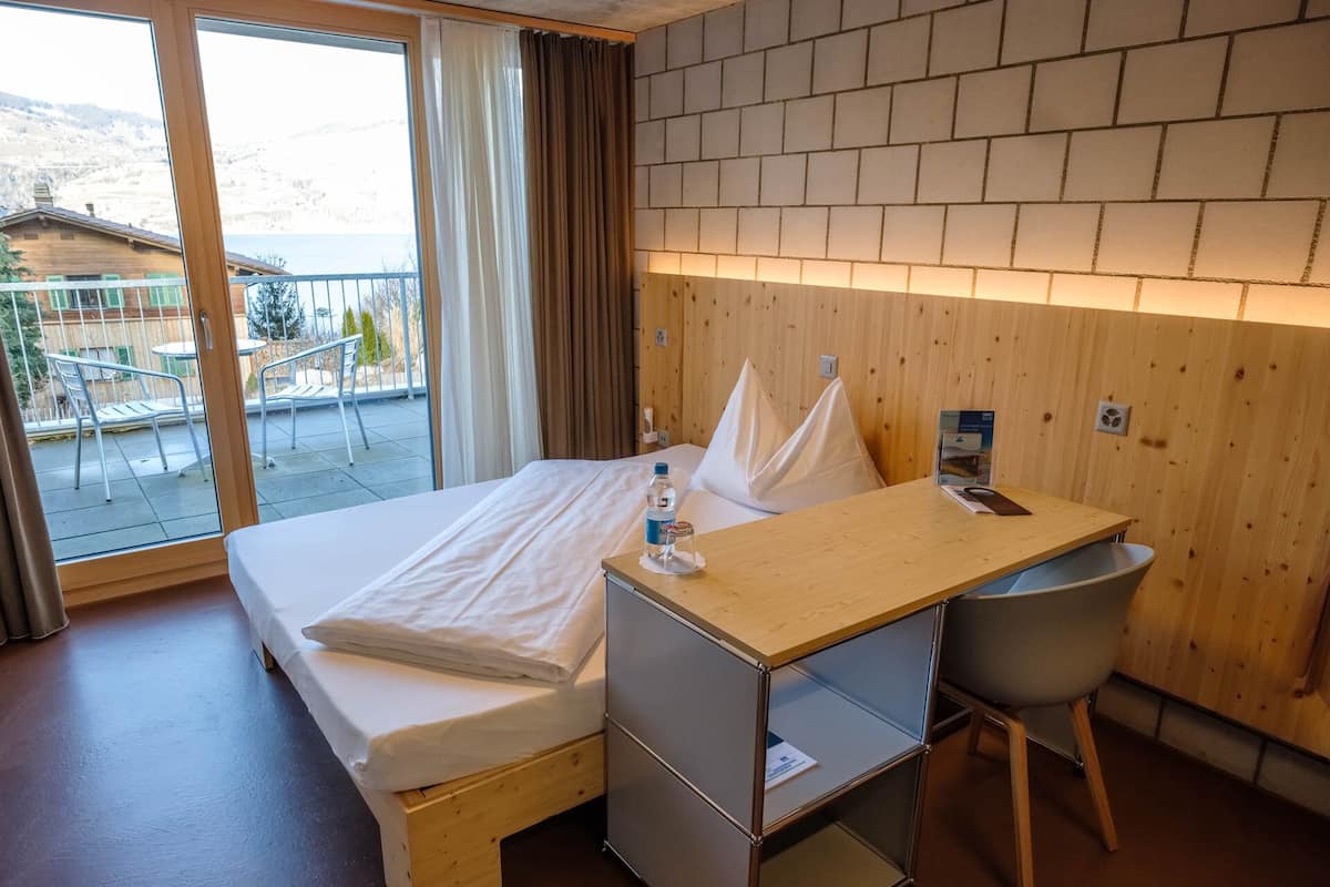 A hotel room with a bed, a wooden desk, a chair, and a view of a balcony with outdoor furniture.
