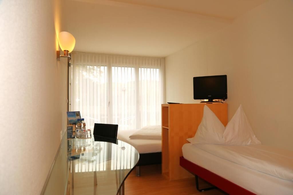 A hotel room with a glass table, a bed, a TV, and a window with blinds.