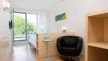 A modern hotel room with a glass door leading to a balcony, a black leather chair, a small round table with a water bottle, and a painting on the wall.