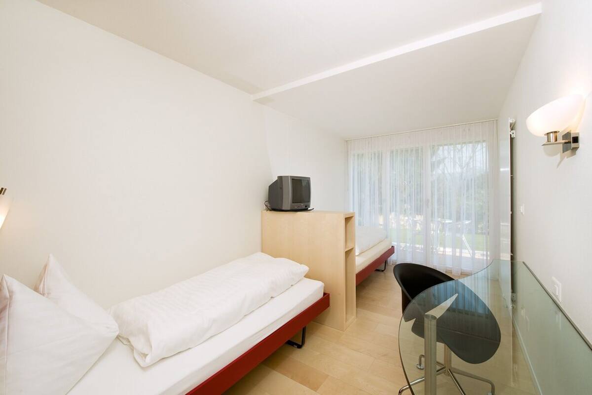 A small, bright room with a single bed, a desk, and a TV.
